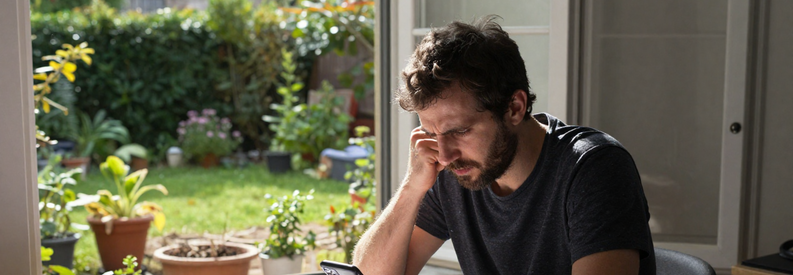 Recherche Jardinier Lyon Frustration Homme frustré cherchant jardinier à Lyon, entouré de papiers et appels téléphoniques, jardin négligé visible par la fenêtre.